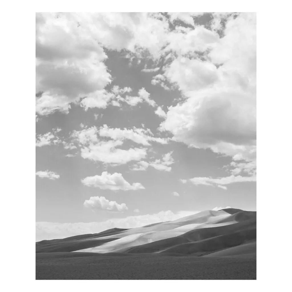 In a black and white photograph, puffy clouds shadow Colorado's Great Sand Dunes, leaving a shaft of light to illuminate the dunes in the center of the frame